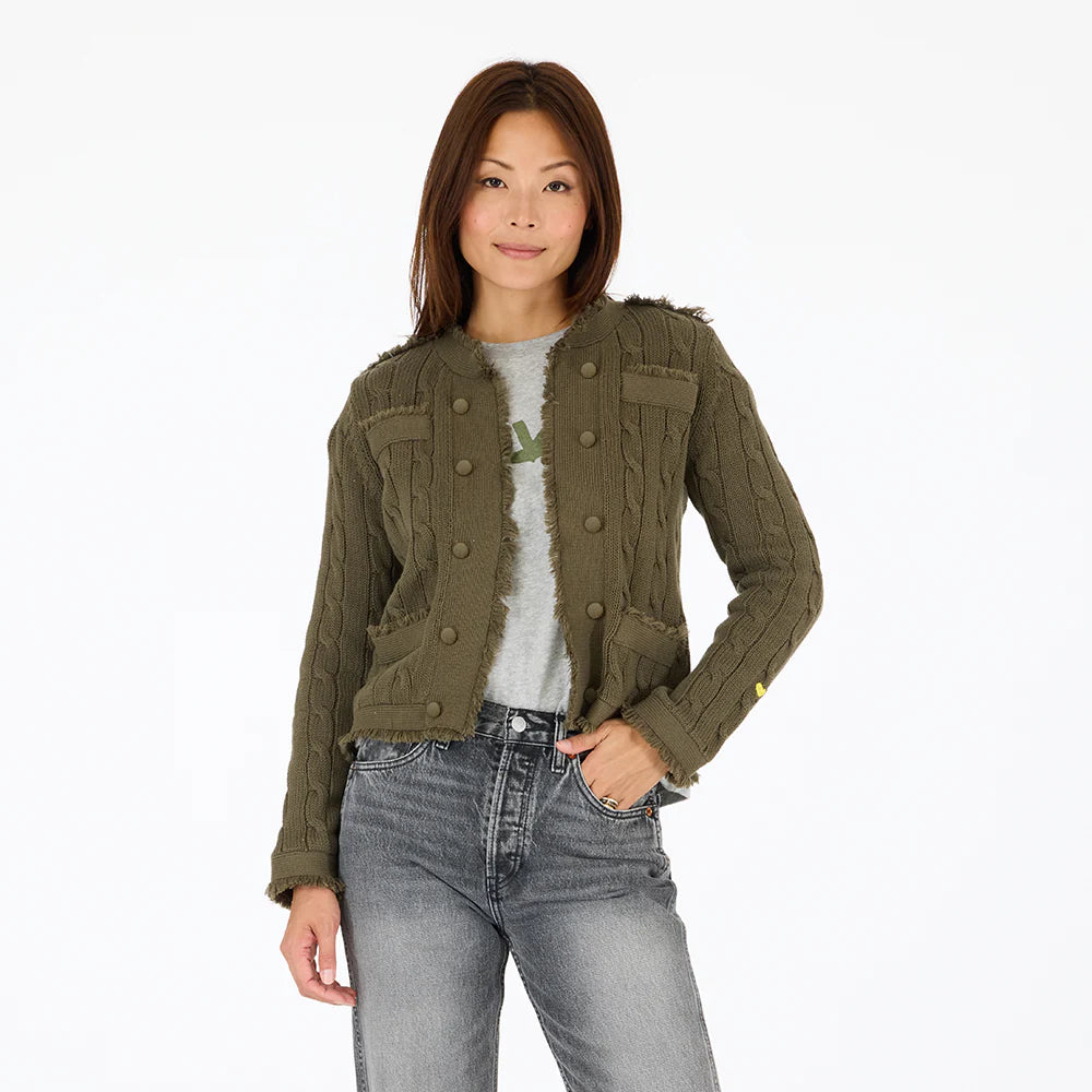 A woman with straight brown hair models the Kerri Rosenthal Beau-Lero Fringe Cardigan in green cotton over a gray shirt and jeans, standing with one hand in her pocket against a plain white background.