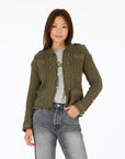 A woman with straight brown hair models the Kerri Rosenthal Beau-Lero Fringe Cardigan in green cotton over a gray shirt and jeans, standing with one hand in her pocket against a plain white background.