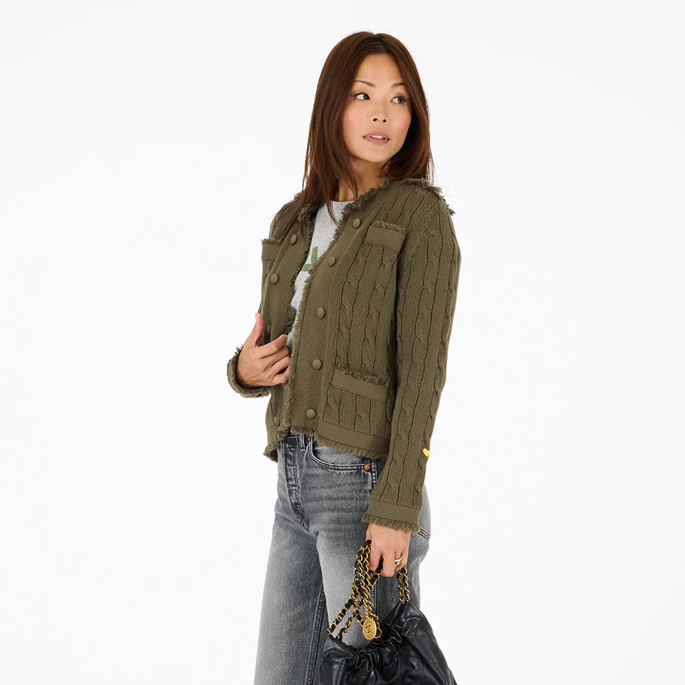 A woman with long brown hair wears the Kerri Rosenthal Beau-Lero Fringe Cardigan in green over a light gray top and gray jeans, holding a black handbag with a gold chain, looking left against a plain white background.