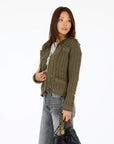 A woman with long brown hair wears the Kerri Rosenthal Beau-Lero Fringe Cardigan in green over a light gray top and gray jeans, holding a black handbag with a gold chain, looking left against a plain white background.