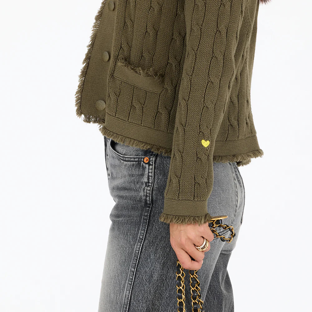 A person wearing the Kerri Rosenthal Beau-Lero Fringe Cardigan in textured olive-green with a crew neck, gray jeans, and a gold chain bag. A small yellow heart accents the 100% cotton cardigan’s sleeve.