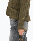 A person wearing the Kerri Rosenthal Beau-Lero Fringe Cardigan in textured olive-green with a crew neck, gray jeans, and a gold chain bag. A small yellow heart accents the 100% cotton cardigan’s sleeve.