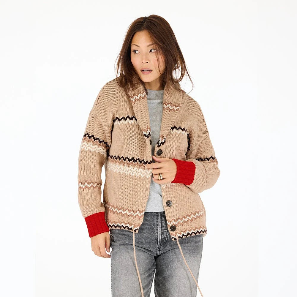 A woman in a beige Kerri Rosenthal Shawl Collar Ker-Isle Cardigan with black and white zigzag patterns and red cuffs, paired with a gray shirt and jeans, stands against a plain white background looking to the side.