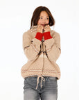 A person wears the Kerri Rosenthal Shawl Collar Ker-Isle Cardigan, beige with red cuffs, against a white background. Smiling, they hold the collar near their face and look to the side, paired with light blue jeans.