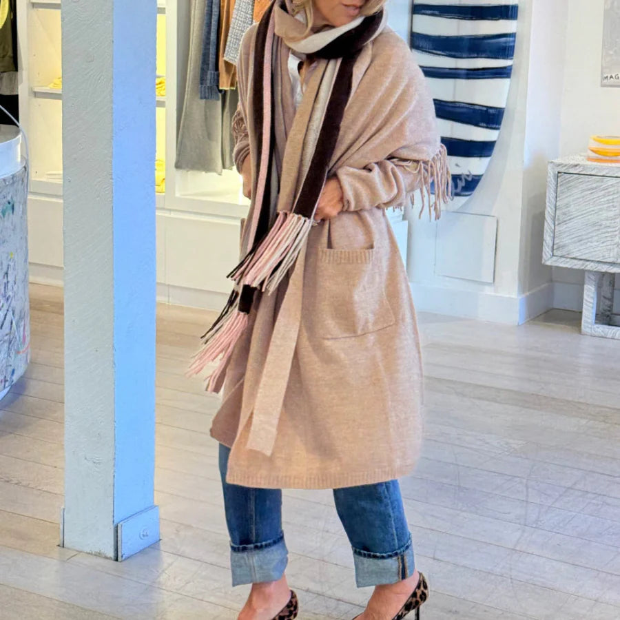 A person wearing the Kerri Rosenthal Cali Fringe Cardigan Coat, cuffed blue jeans, a multi-colored scarf partially covering their face, and leopard print shoes stands indoors near a blue pillar.