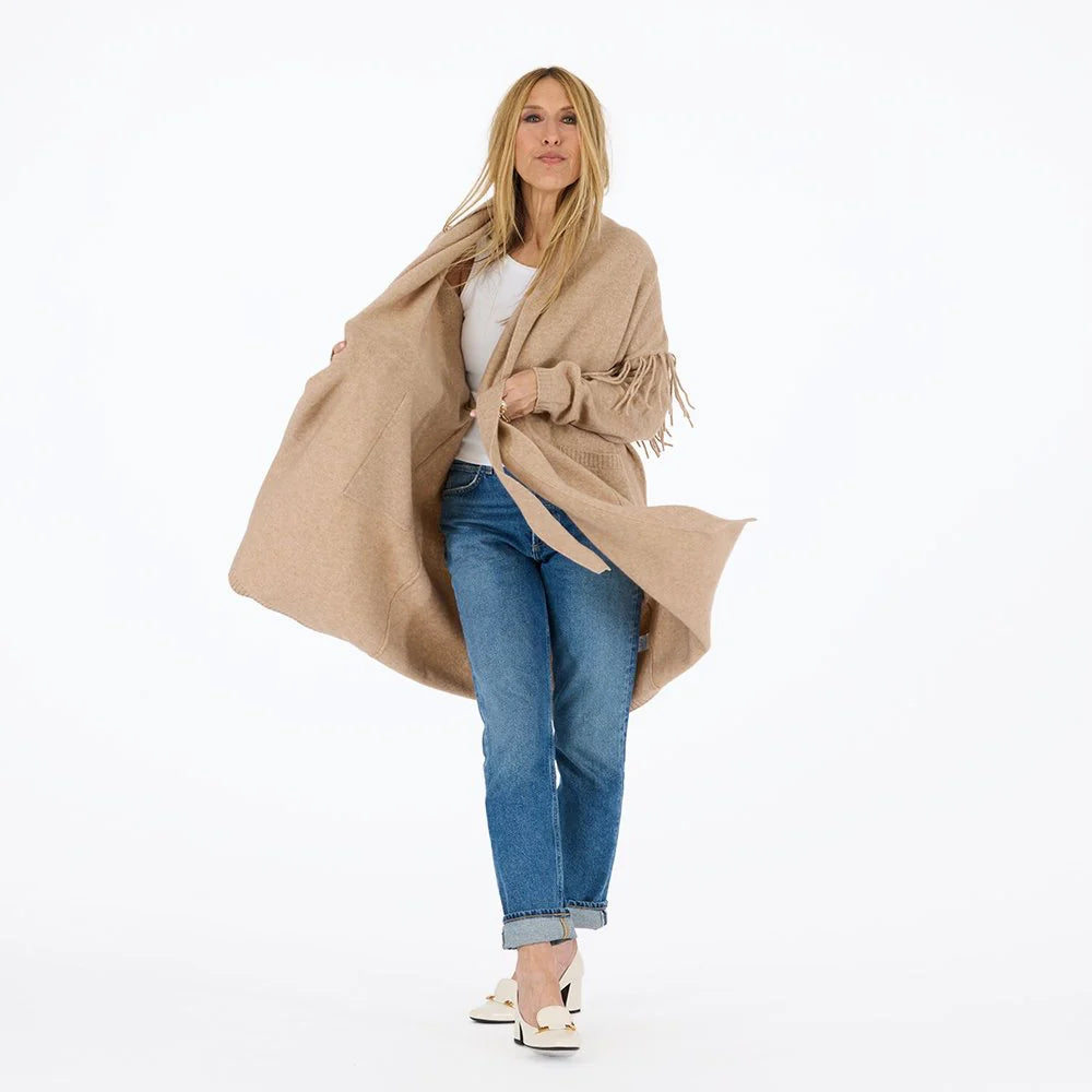 A woman with long blonde hair stands against a white background, wearing the Kerri Rosenthal Cali Fringe Cardigan Coat over a white top, blue cuffed jeans, and white heels. She is looking slightly to the side.