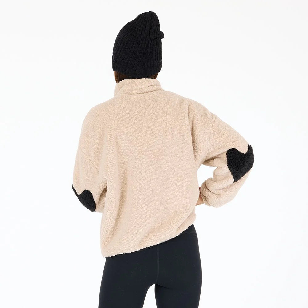 A person wearing the Kerri Rosenthal Coze Magoze—a beige cotton fleece jacket with heart elbows—black leggings, and a black knit beanie stands facing away against a plain white background.