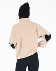 A person wearing the Kerri Rosenthal Coze Magoze—a beige cotton fleece jacket with heart elbows—black leggings, and a black knit beanie stands facing away against a plain white background.