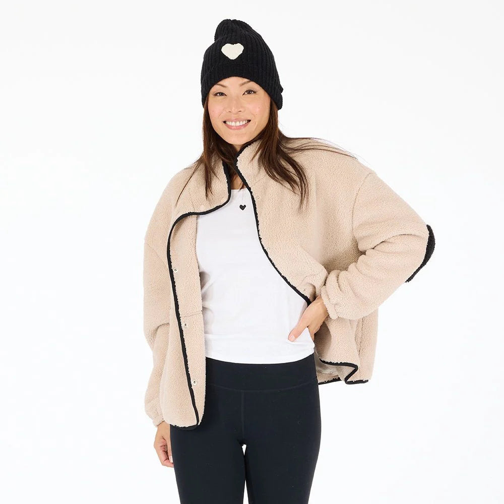 A woman smiles in a beige Coze Magoze jacket by Kerri Rosenthal with heart elbows, paired with a white shirt, black leggings, and a black knit hat with a heart, standing against a white background.