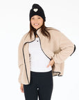 A woman smiles in a beige Coze Magoze jacket by Kerri Rosenthal with heart elbows, paired with a white shirt, black leggings, and a black knit hat with a heart, standing against a white background.
