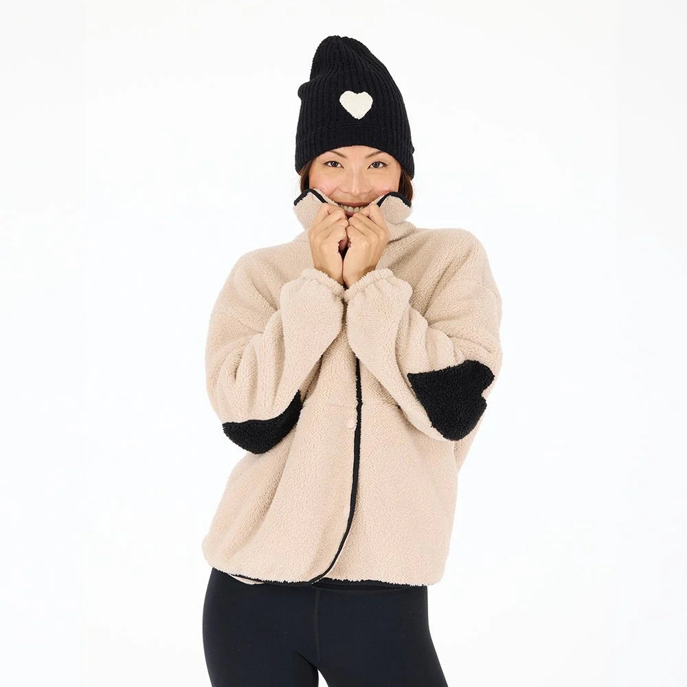 A person smiles against a plain white background, holding up the collar of their beige Kerri Rosenthal Coze Magoze jacket with black heart elbows and wearing a black beanie with a white heart.