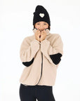 A person smiles against a plain white background, holding up the collar of their beige Kerri Rosenthal Coze Magoze jacket with black heart elbows and wearing a black beanie with a white heart.