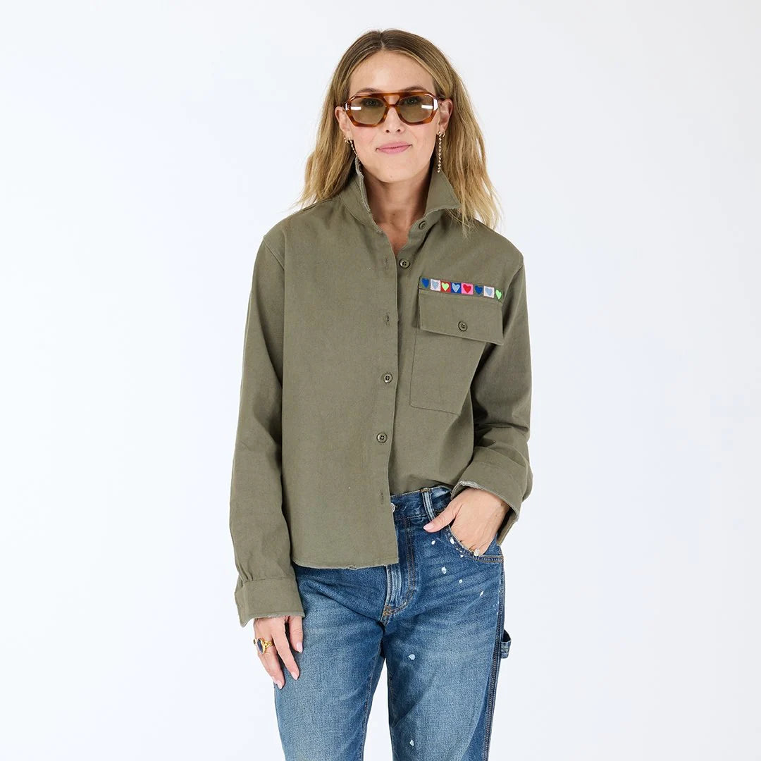 A woman in the Kerri Rosenthal Pia Love Brigade Shirt, featuring colorful pins on the olive green pocket and heart patch elbows, paired with blue jeans and sunglasses, stands against a plain white background.