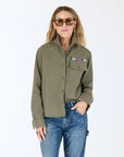 A woman in the Kerri Rosenthal Pia Love Brigade Shirt, featuring colorful pins on the olive green pocket and heart patch elbows, paired with blue jeans and sunglasses, stands against a plain white background.