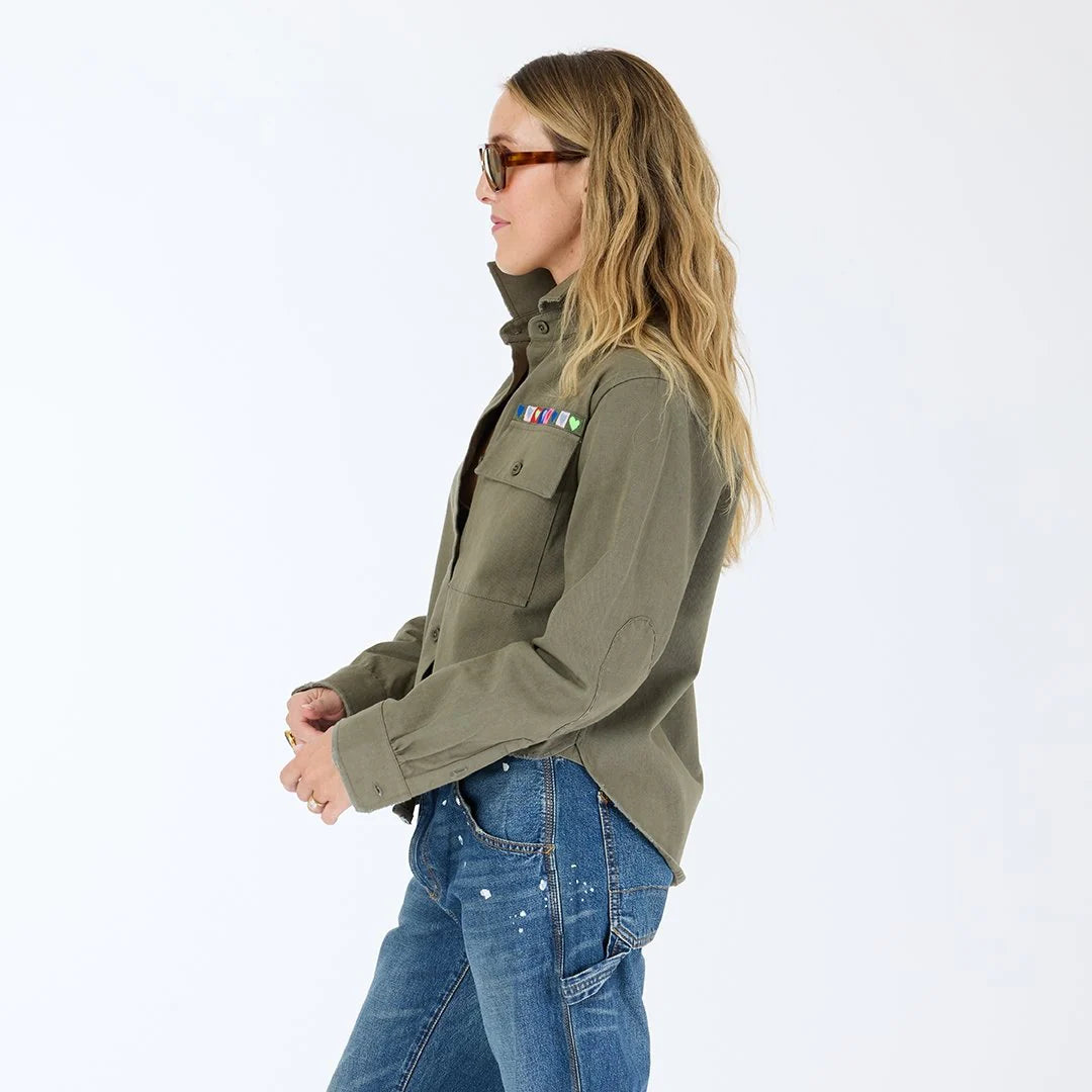 A woman with long, wavy blonde hair wears sunglasses, blue jeans with paint splatters, and the Kerri Rosenthal Pia Love Brigade Shirt featuring colorful pocket patches and heart patch elbows, standing against a plain white background.