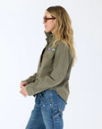A woman with long, wavy blonde hair wears sunglasses, blue jeans with paint splatters, and the Kerri Rosenthal Pia Love Brigade Shirt featuring colorful pocket patches and heart patch elbows, standing against a plain white background.