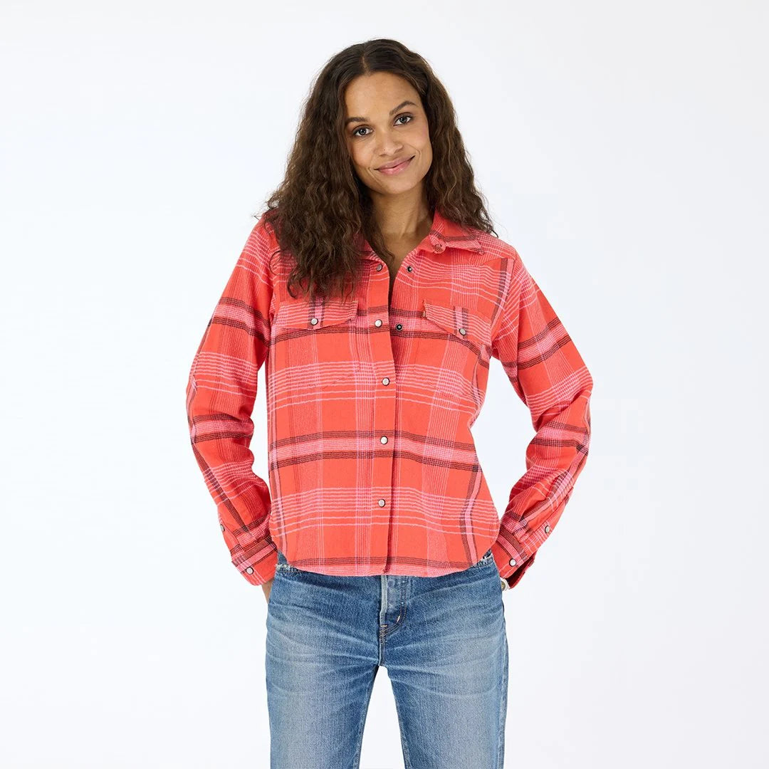 A woman with long curly brown hair smiles with her hands in her pockets, wearing the Kerri Rosenthal Pia Western Flannel and blue jeans against a plain white background.
