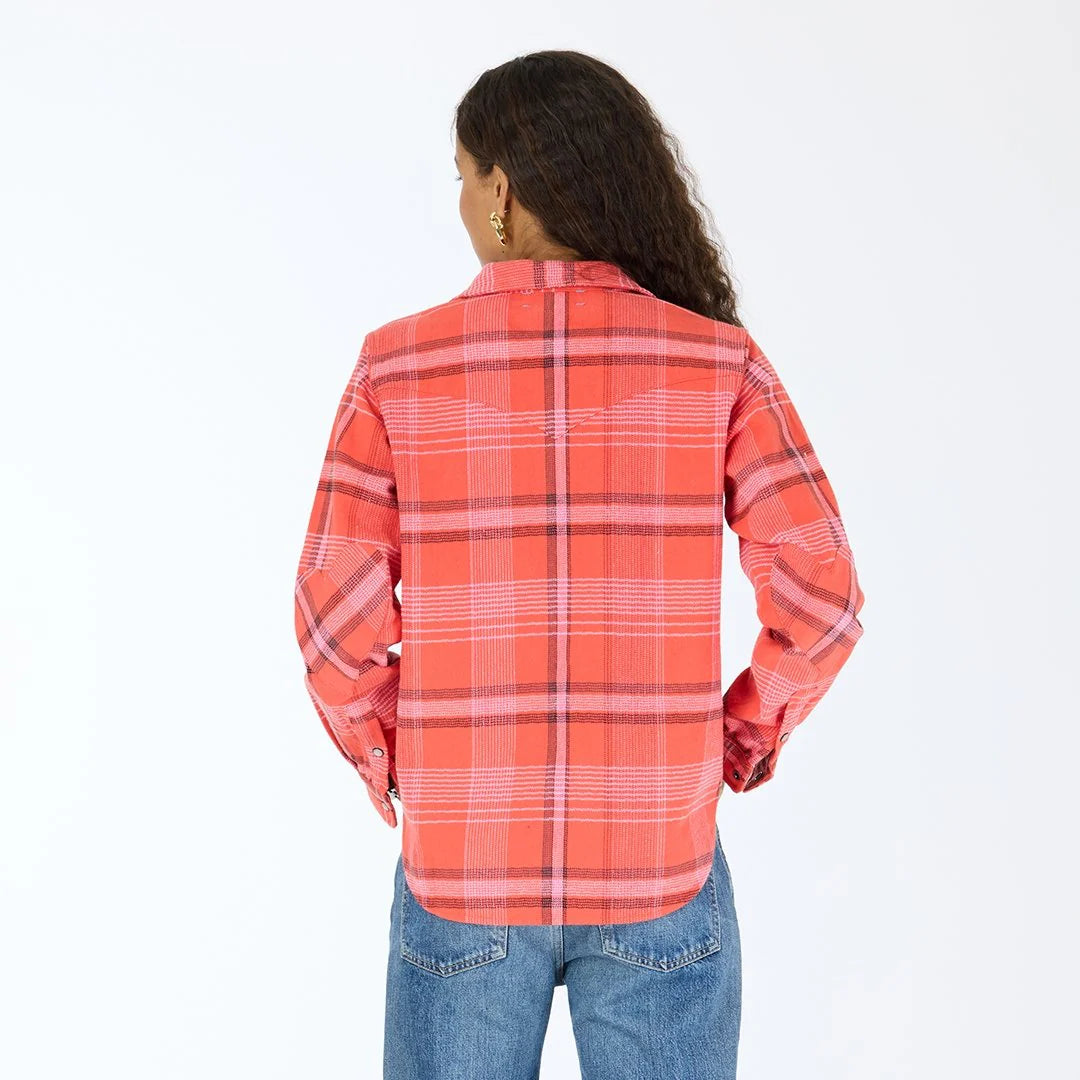 A person with long curly hair, wearing the Kerri Rosenthal Pia Western Flannel in red and pink and blue jeans, stands facing away from the camera against a plain white background.