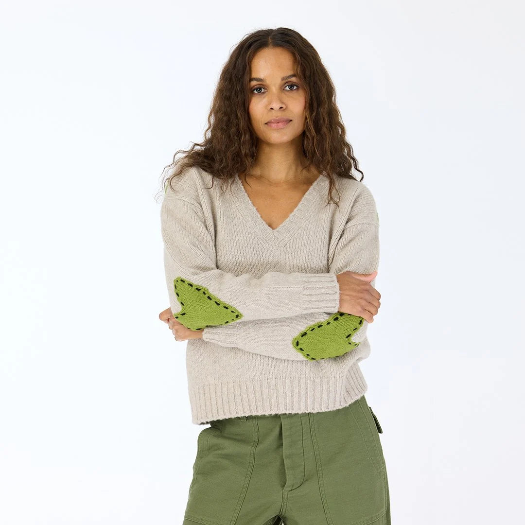 A woman with long curly hair, arms crossed, models the Kerri Rosenthal Frenchy Sweater—beige cashmere with green heart patch elbows—and green pants against a plain white background.