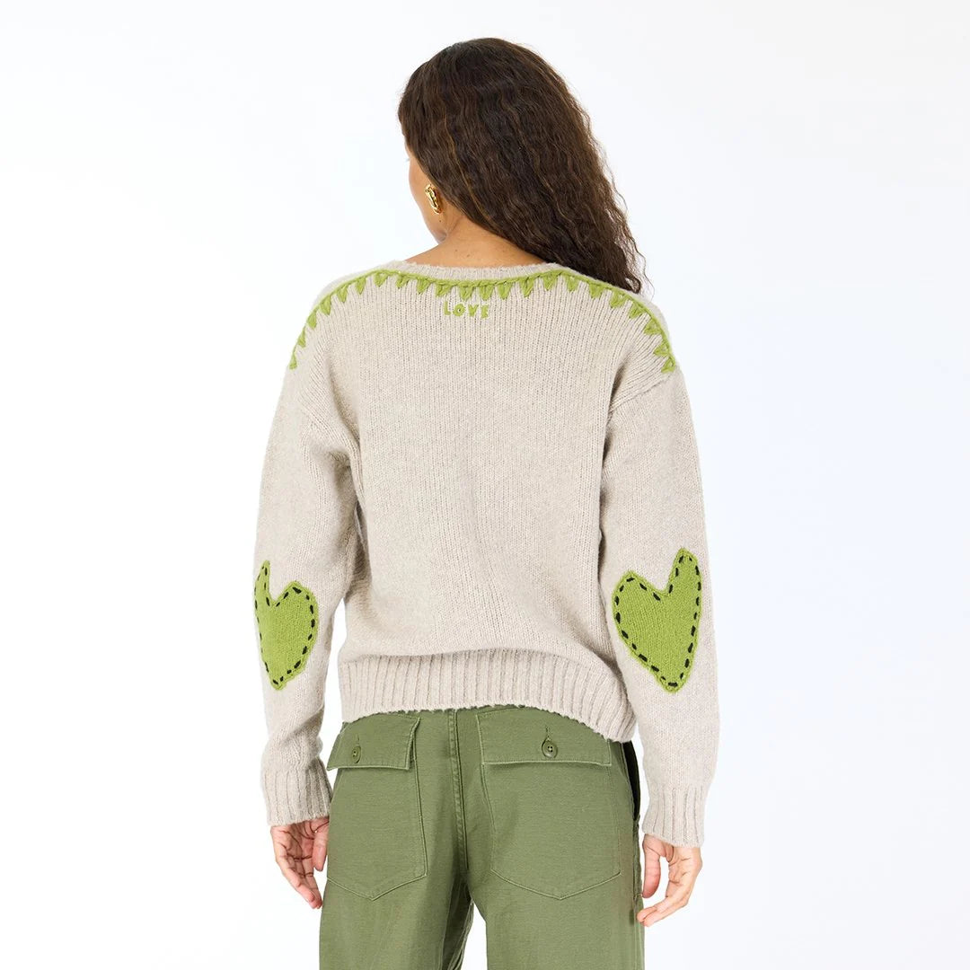 A person with long curly hair is seen from the back wearing the Kerri Rosenthal Frenchy Sweater in beige, featuring heart patch elbows and green whipstitch details, paired with olive green pants against a plain white background.