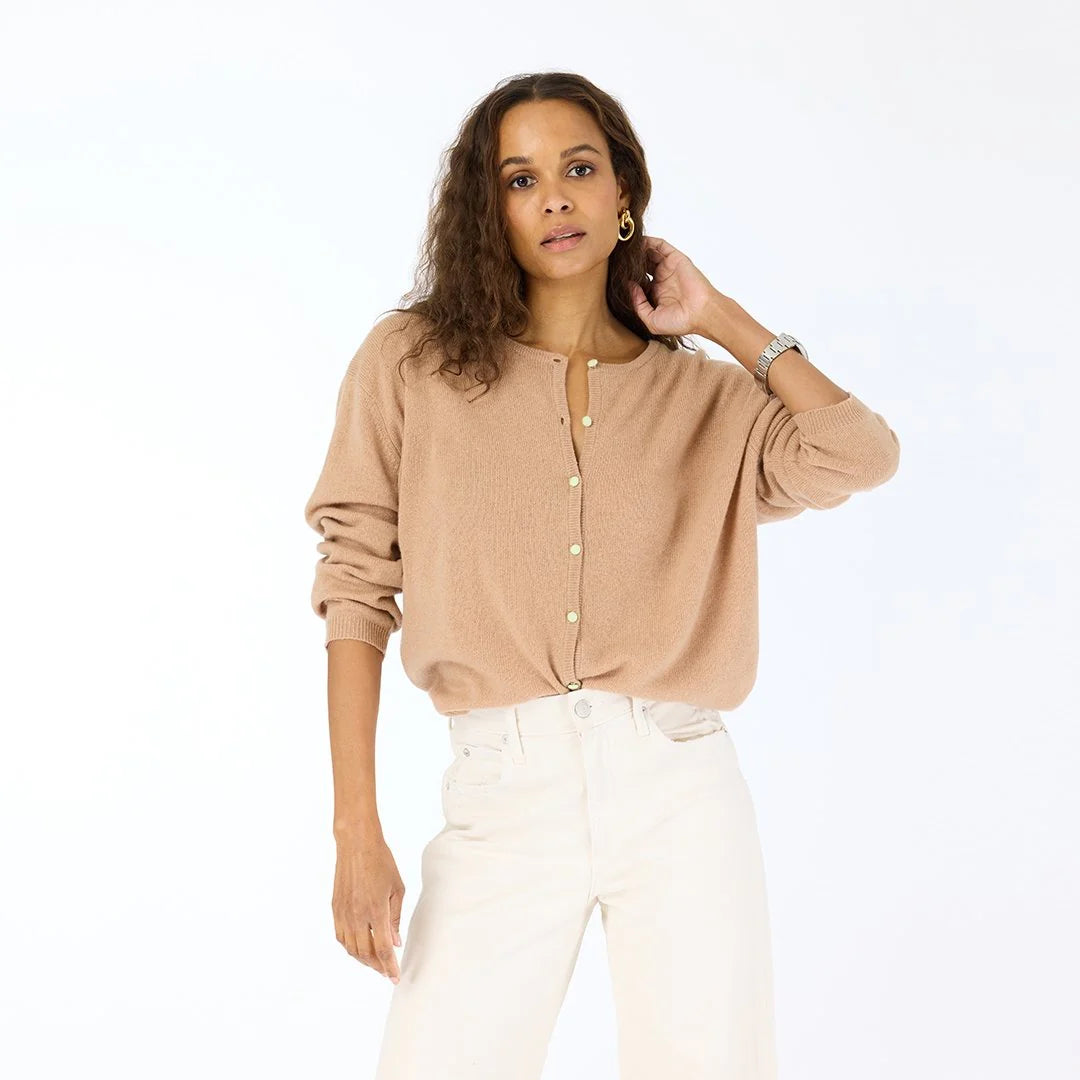 A woman with wavy brown hair wears the beige Kerri Rosenthal Bassett Cardigan and white high-waisted pants, standing against a plain white background with her left hand touching her head.