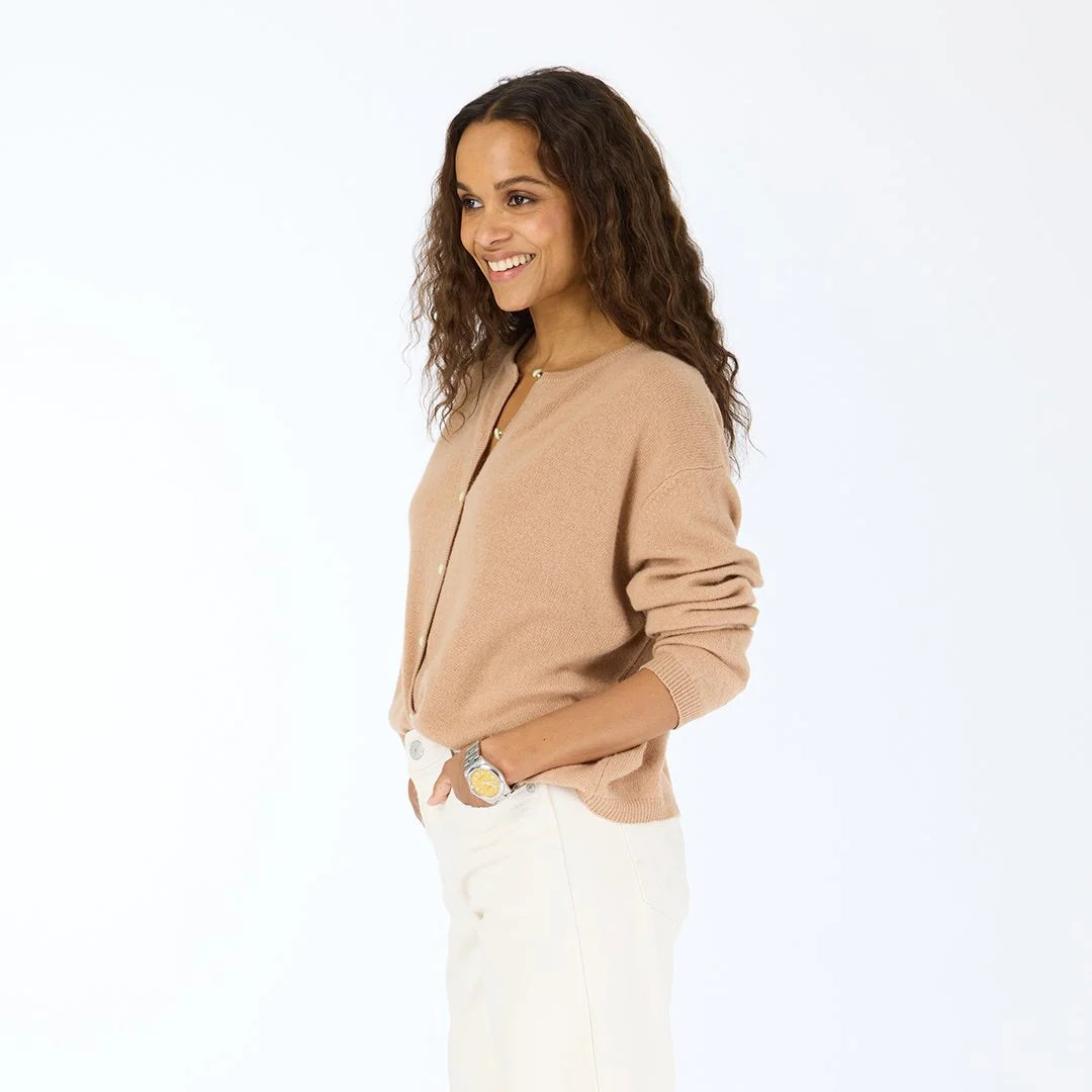 A woman with long, curly hair smiles in profile, wearing the Kerri Rosenthal Bassett Cardigan, white pants, and a yellow watch. Her hands are in her pockets as she poses against a plain white background.