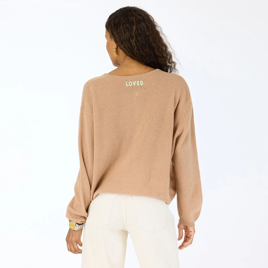 A woman with wavy hair, facing away, wears a light brown Kerri Rosenthal Bassett Cardigan in featherweight cashmere with "LOVED" on the back neckline and light pants, standing against a plain white background.