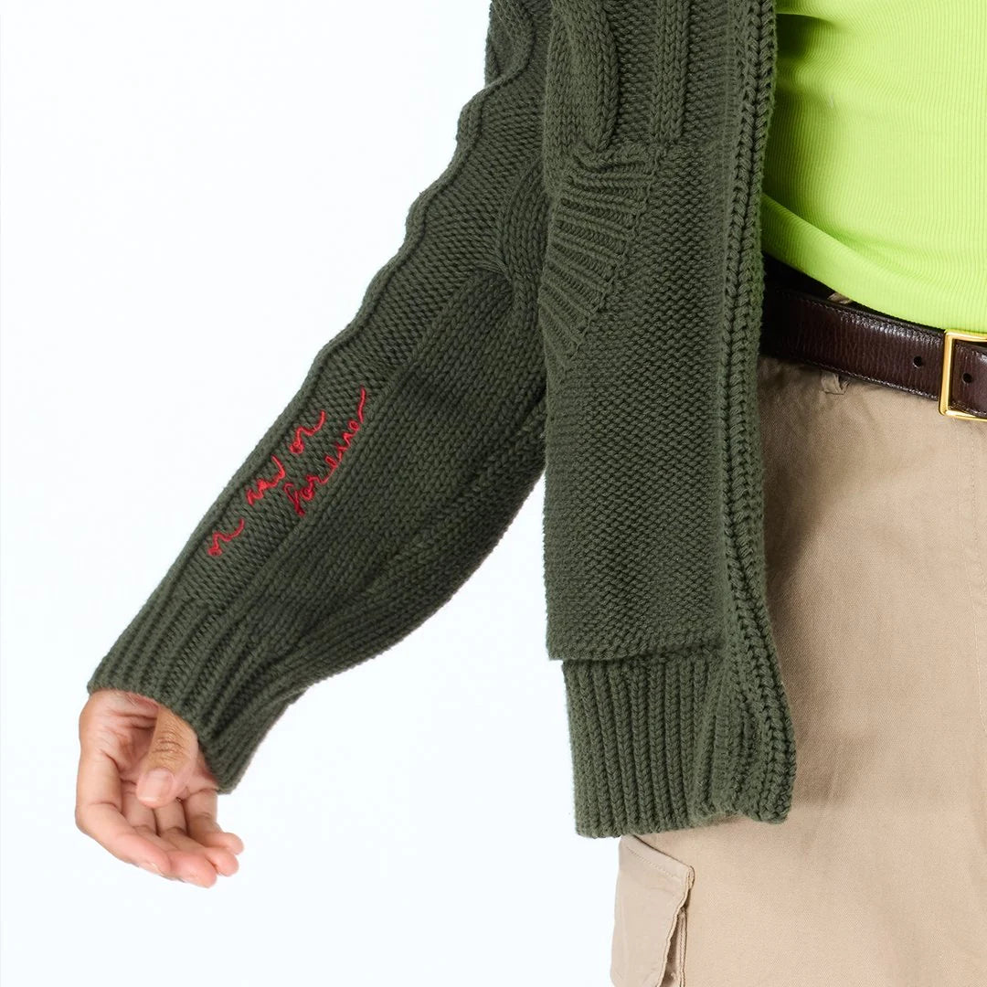 A person wears the Kerri Rosenthal Shawl Collar Helen Cardigan in green over a neon green shirt and khaki pants. The cardigan's sleeve features red cursive embroidery, with their relaxed hand visible.