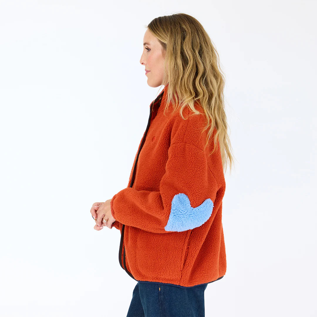 A woman with long wavy hair wears the cozy Kerri Rosenthal Coze Magoze cotton fleece jacket in orange with a blue heart elbow patch, standing sideways against a plain white background.