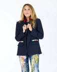 A woman with long blonde hair smiles, wearing the Kerri Rosenthal Workday Love Brigade Blazer with colorful artful details and pocket accents, paired with paint-splattered jeans, standing against a plain white background.