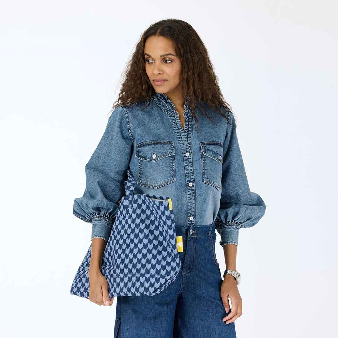 A woman in a denim shirt and blue jeans holds the Kerri Rosenthal KR Heart in Check Tote, a large blue cotton canvas bag with drippy hearts. She has long curly hair and looks left, standing against a plain white background.