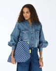 A woman in a denim shirt and blue jeans holds the Kerri Rosenthal KR Heart in Check Tote, a large blue cotton canvas bag with drippy hearts. She has long curly hair and looks left, standing against a plain white background.