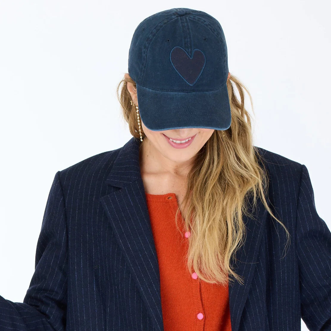 A woman with long blonde hair smiles while looking down, wearing a navy KR Imperfect Heart Hat by Kerri Rosenthal, featuring an embroidered heart patch, paired with a dark pinstripe blazer and orange cardigan against a white background.