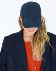 A woman with long blonde hair smiles while looking down, wearing a navy KR Imperfect Heart Hat by Kerri Rosenthal, featuring an embroidered heart patch, paired with a dark pinstripe blazer and orange cardigan against a white background.