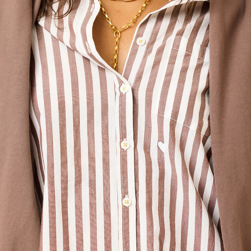 Close-up of a person wearing the Kerri Rosenthal Mia Stripe Shirt—brown and white cotton stripes with a small white heart embroidered on the pocket—styled with a gold chain necklace and a brown jacket over the shoulders.