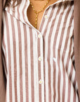 Close-up of a person wearing the Kerri Rosenthal Mia Stripe Shirt—brown and white cotton stripes with a small white heart embroidered on the pocket—styled with a gold chain necklace and a brown jacket over the shoulders.