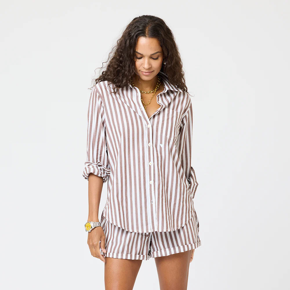 A woman with long curly hair wears the Kerri Rosenthal Mia Stripe Shirt, a brown and white striped cotton button-down with rolled sleeves and matching shorts, accessorized with gold jewelry, standing against a plain white background.