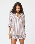 A woman with long curly hair wears the Kerri Rosenthal Mia Stripe Shirt, a brown and white striped cotton button-down with rolled sleeves and matching shorts, accessorized with gold jewelry, standing against a plain white background.