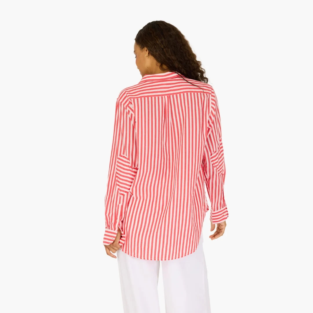 A woman with curly hair wears the Kerri Rosenthal Mia Stripe Shirt—a long-sleeve red and white striped cotton shirt with heart embroidery—paired with white pants, standing with her back to the camera against a plain white background.