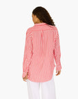 A woman with curly hair wears the Kerri Rosenthal Mia Stripe Shirt—a long-sleeve red and white striped cotton shirt with heart embroidery—paired with white pants, standing with her back to the camera against a plain white background.