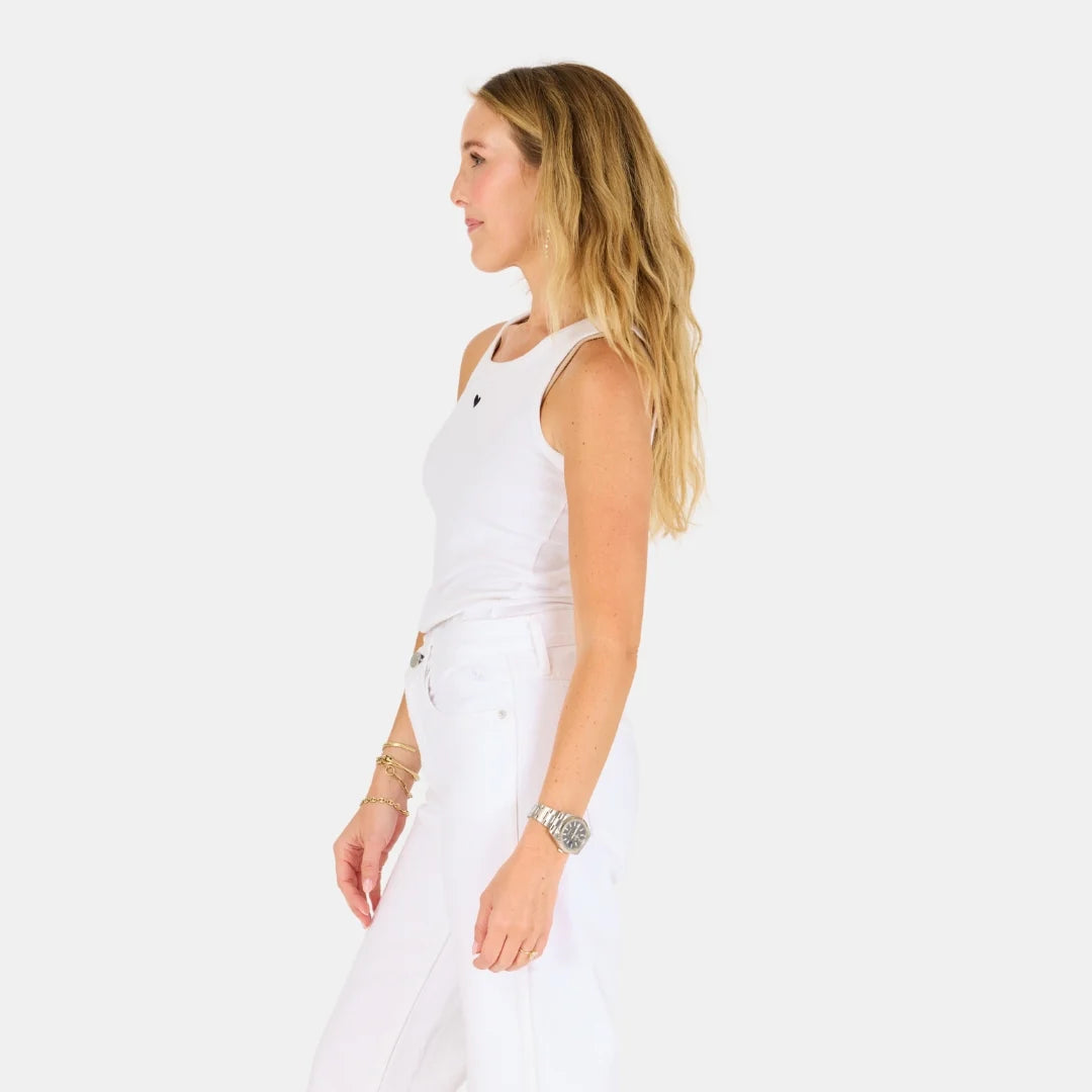 A woman with long blonde hair stands in profile against a white background, wearing the KR Tank by Kerri Rosenthal and white pants, accessorized with bracelets and a watch, showcasing effortless style.