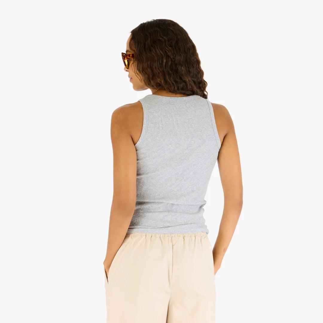 A person with long, wavy hair wears sunglasses and the Kerri Rosenthal KR Tank—a black cotton top with an embroidered heart—paired with beige pants. Shown from the back, hands in pockets, against a plain white background.