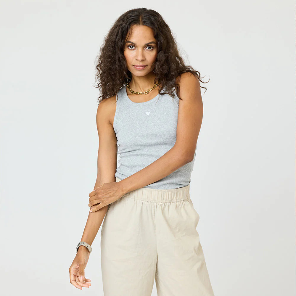 A woman with long curly hair wears the Kerri Rosenthal KR Tank in gray with light beige pants, a gold necklace, and a watch. She stands against a plain light background, looking at the camera and touching her arm.