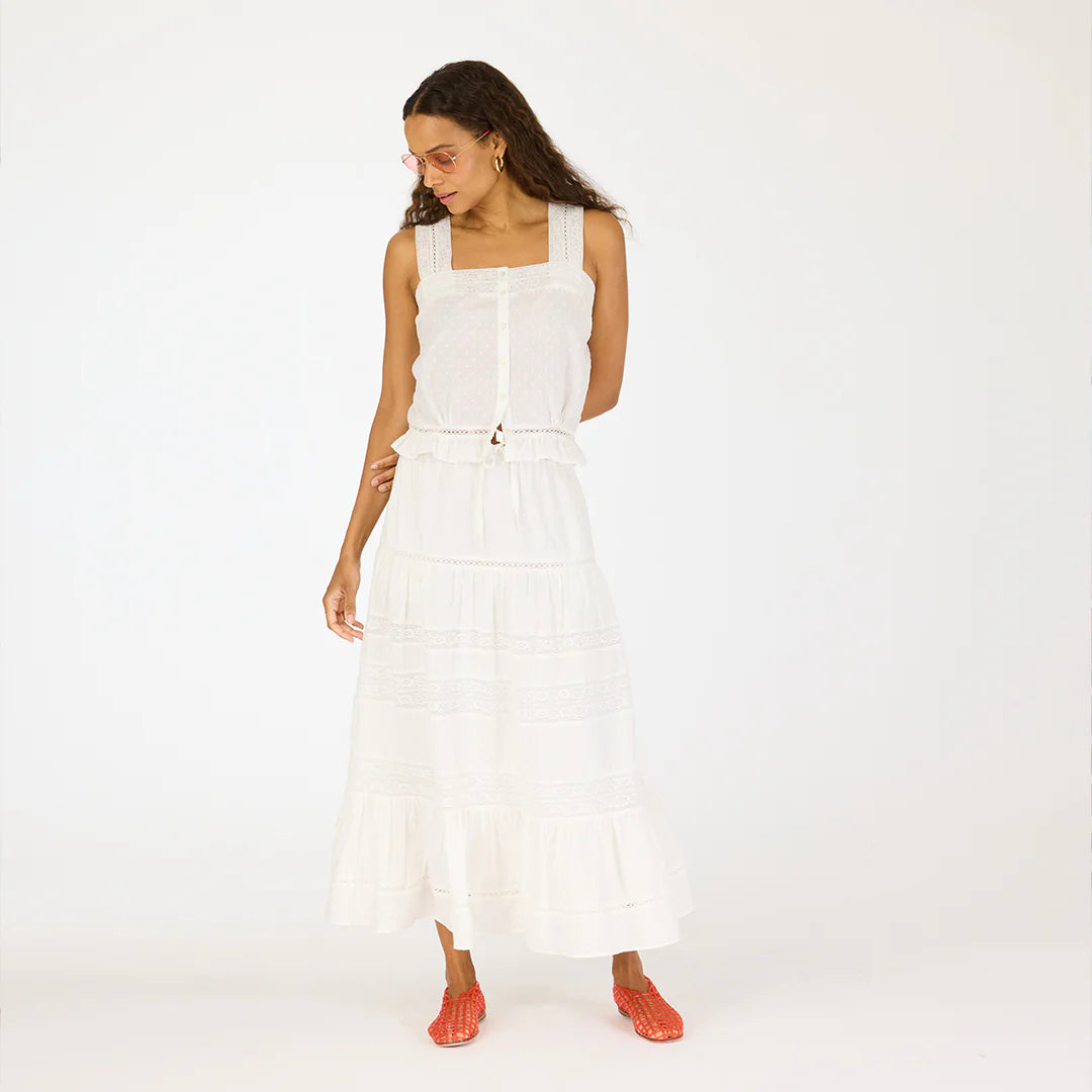 A woman with long curly hair and glasses stands against a white background wearing a sleeveless white top, orange woven shoes, and the Kerri Rosenthal Vacance Skirt featuring ruffles and subtle lace trim, looking slightly down to her right.