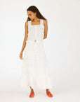 A woman with long curly hair and glasses stands against a white background wearing a sleeveless white top, orange woven shoes, and the Kerri Rosenthal Vacance Skirt featuring ruffles and subtle lace trim, looking slightly down to her right.