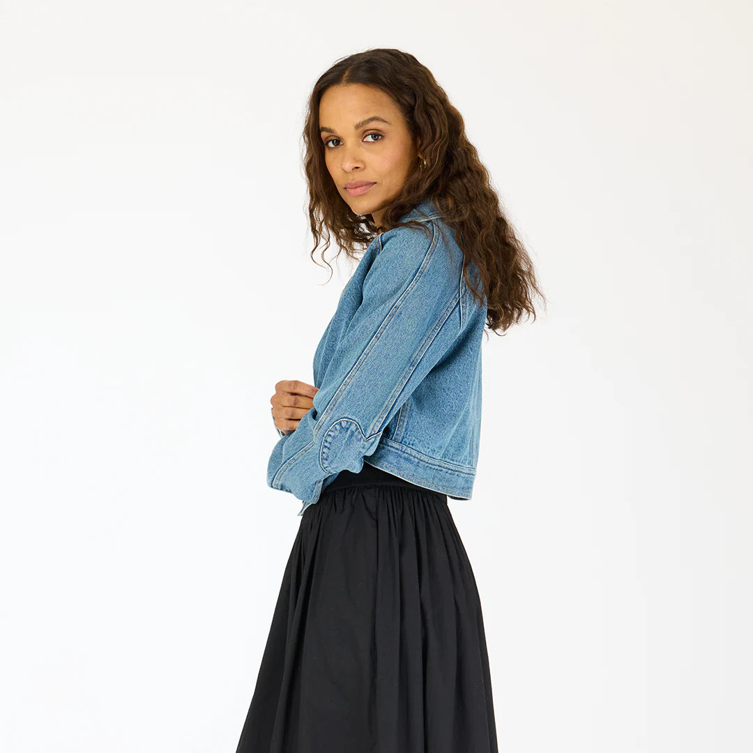 A woman with long curly hair models the Kerri Rosenthal Denim Jacket featuring heart elbow patches, paired with a black skirt. She stands sideways, looking at the camera against a plain white background.