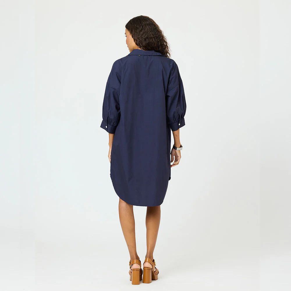 A woman with curly hair faces away, wearing the Kerri Rosenthal Gigi Denim Dress—a loose-fitting, navy blue, knee-length cotton shirt dress with three-quarter sleeves—and tan high-heeled sandals against a plain white background.