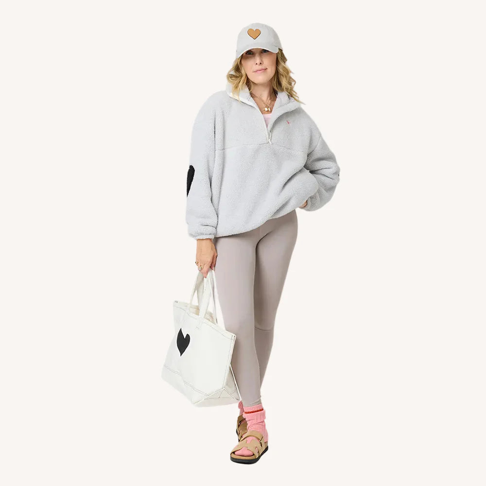 A woman wears a light gray fleece pullover, beige leggings, tan sandals with pink socks, and a Kerri Rosenthal KR Leather Heart Hat. She carries a white tote bag; both showcase the KR Leather Heart Hat design.