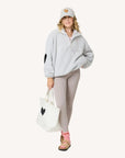 A woman wears a light gray fleece pullover, beige leggings, tan sandals with pink socks, and a Kerri Rosenthal KR Leather Heart Hat. She carries a white tote bag; both showcase the KR Leather Heart Hat design.