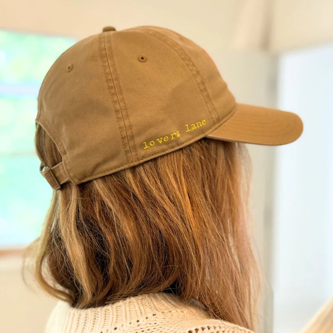 A person with light brown hair wears the Kerri Rosenthal Baseball Hat Heart Patch in tan, featuring "lovers lane" and a yellow heart embroidered on the side. The trendy hat is worn indoors, viewed from behind.